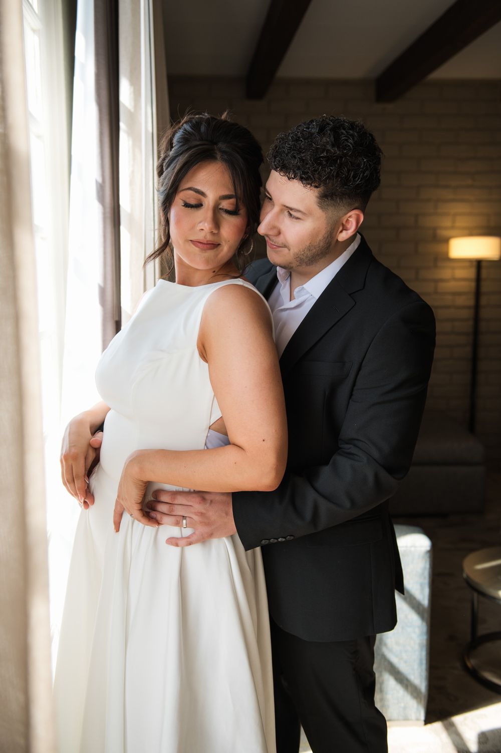 A gentle embrace near a window with natural light streaming in during a wedding photoshoot.