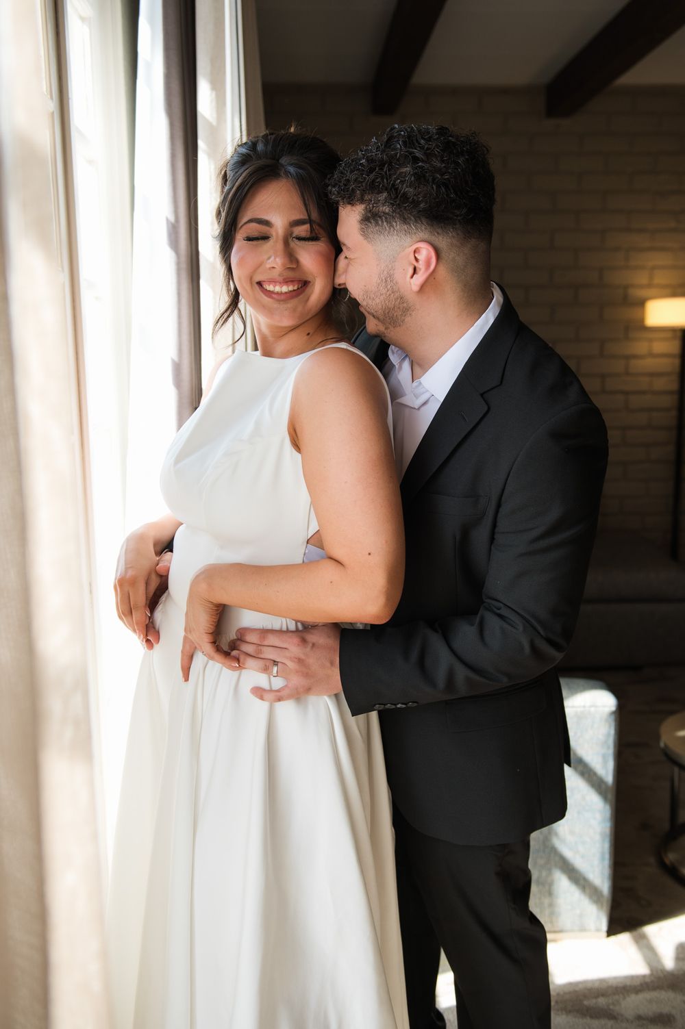 Candid wedding photo capturing joyful laughter between newlyweds by a sunlit window.