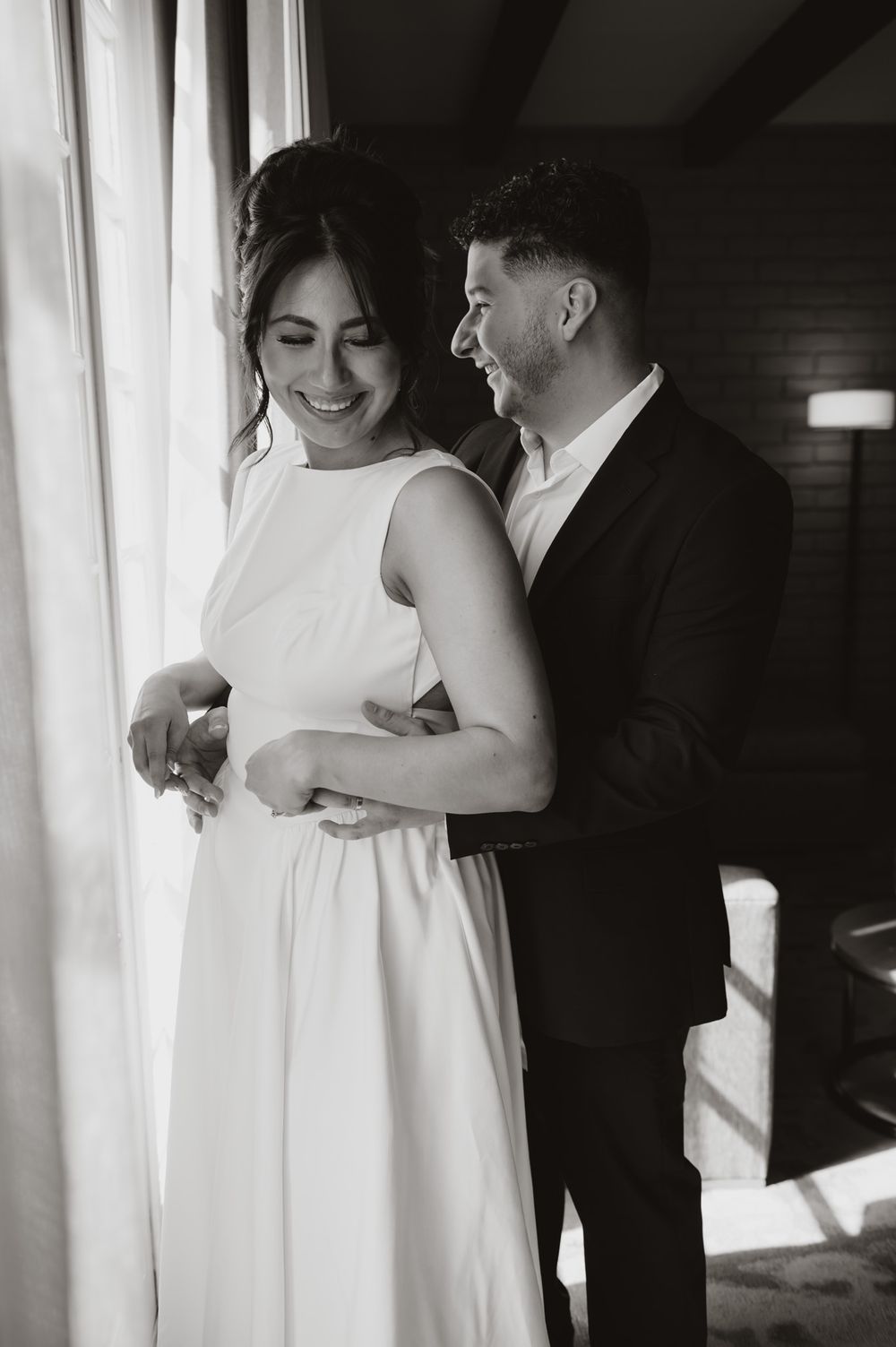 Black and white wedding photo showing a loving embrace near a bright window.