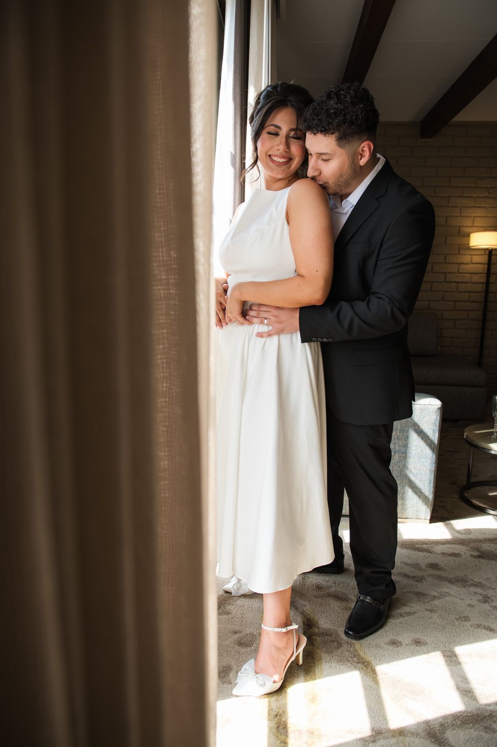 Sunlight streams through window as wedding couple shares intimate moment in formal attire.
