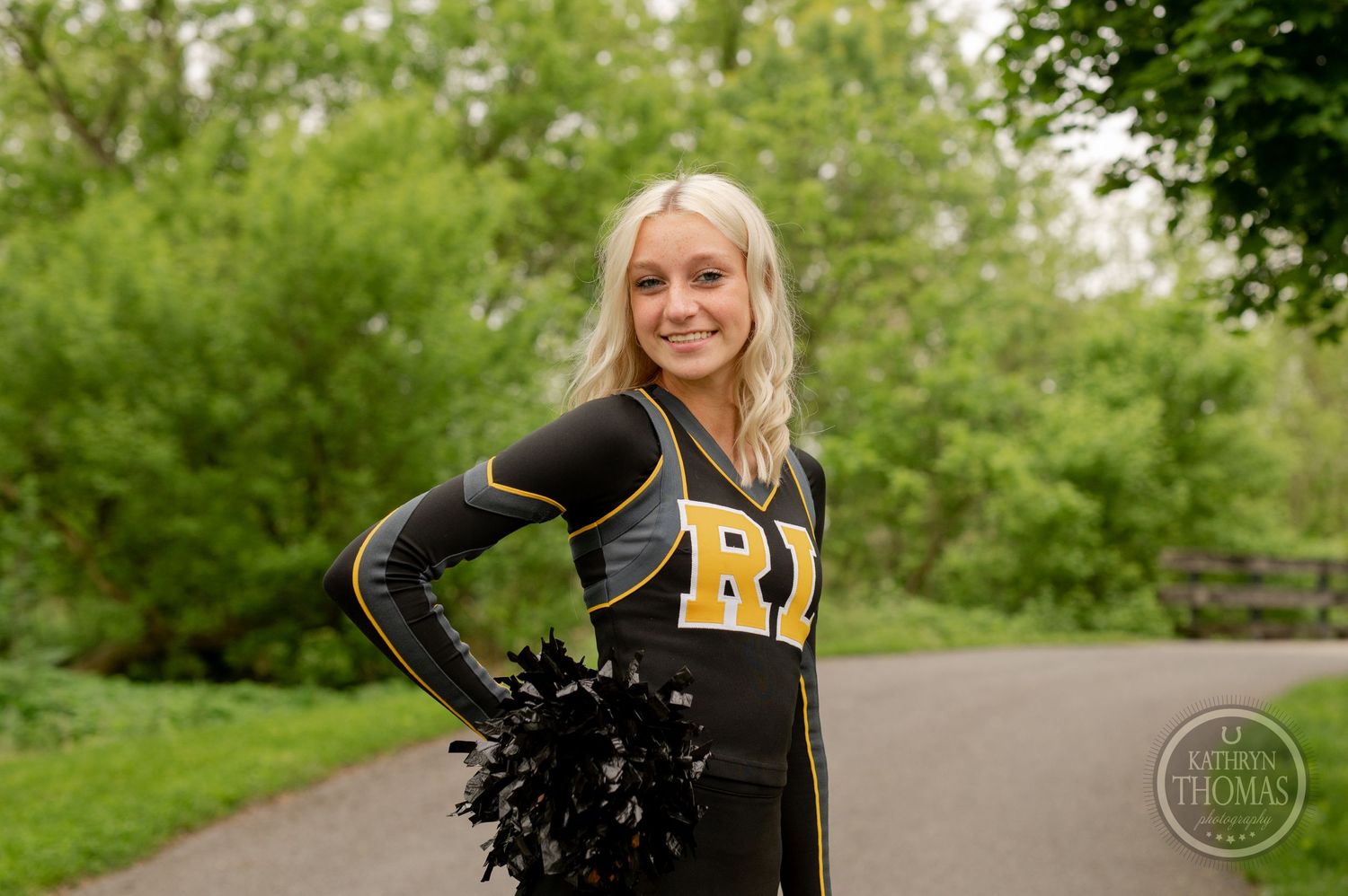 Cheerleader in black uniform with yellow RL letters poses with pom poms in a park setting.