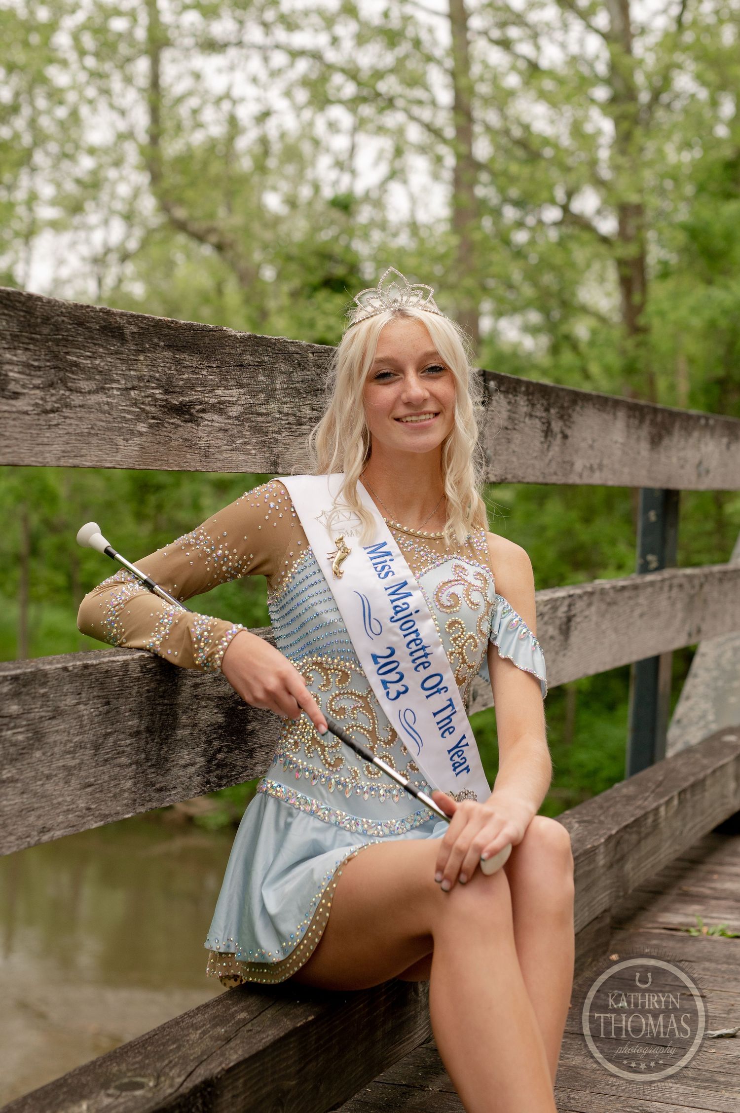 Beauty queen with crown and sash poses on a wooden bridge in an embellished dress.