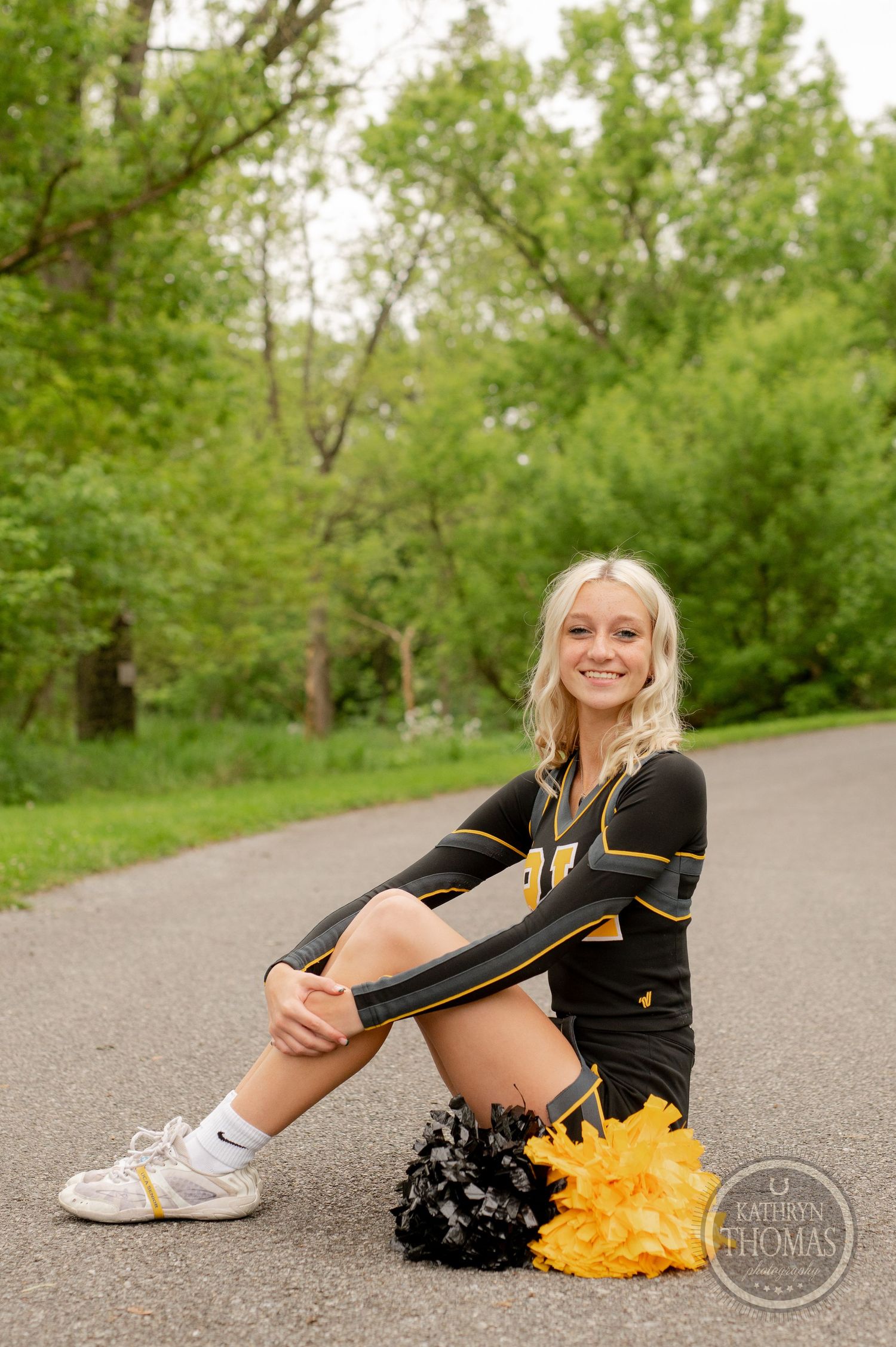 Athletic performer in black and gold uniform sits on path with pom poms among spring greenery.