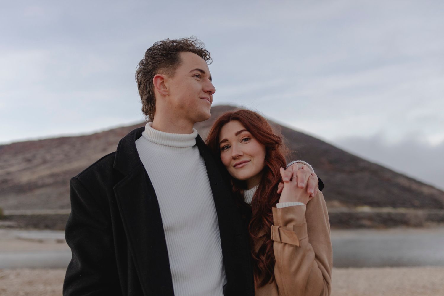 Couple stands in front of mountains. He wraps his arm around her and she holds that hand.