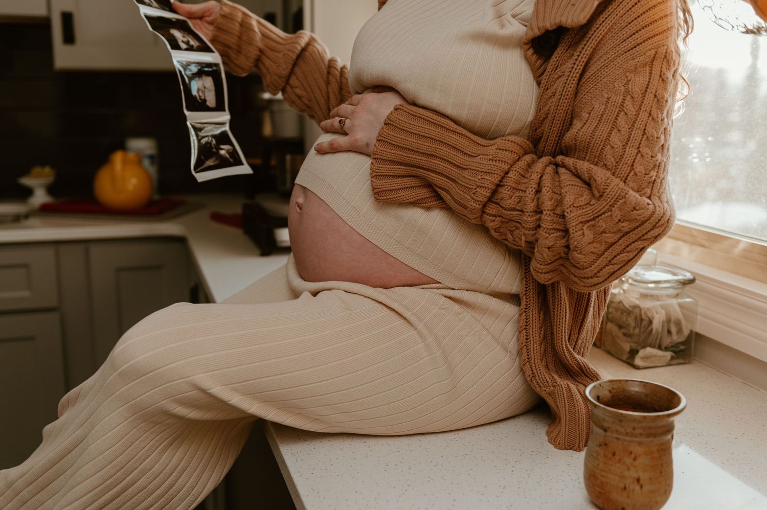 Person in cozy autumn attire sits on counter holding ultrasound photos with coffee cup nearby.