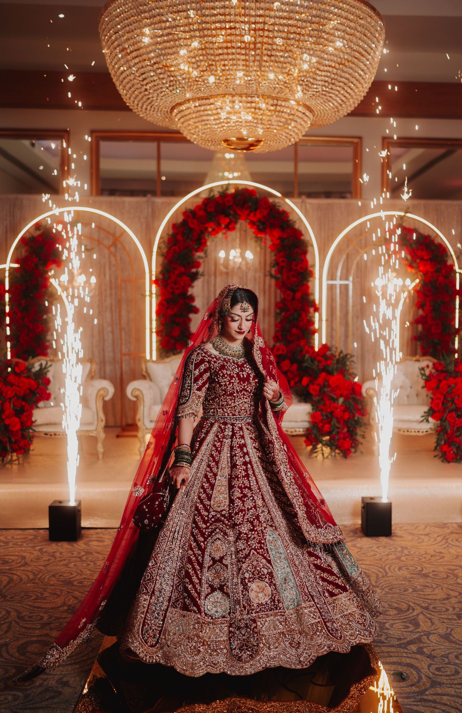 Ornate red bridal lehenga under crystal chandelier with sparkler effects and floral arches.