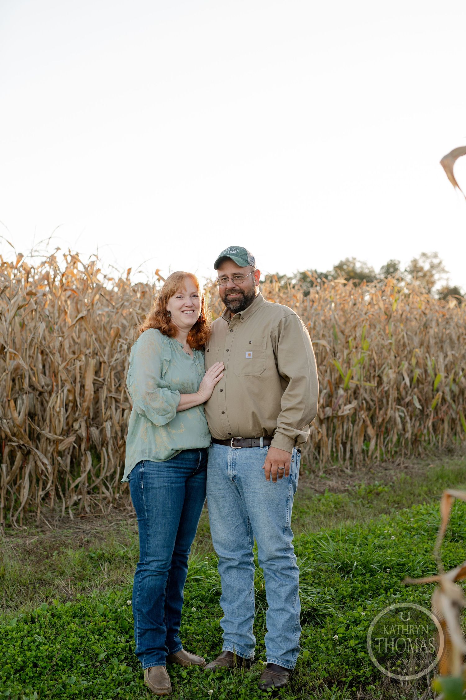 A couple stands together at the edge of a cornfield during sunset.