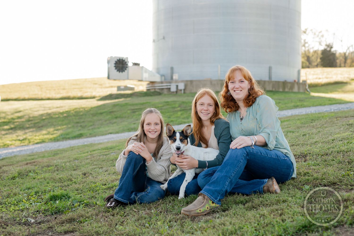 Three people and a spotted dog pose together on grassy hill with large grain silo behind them.