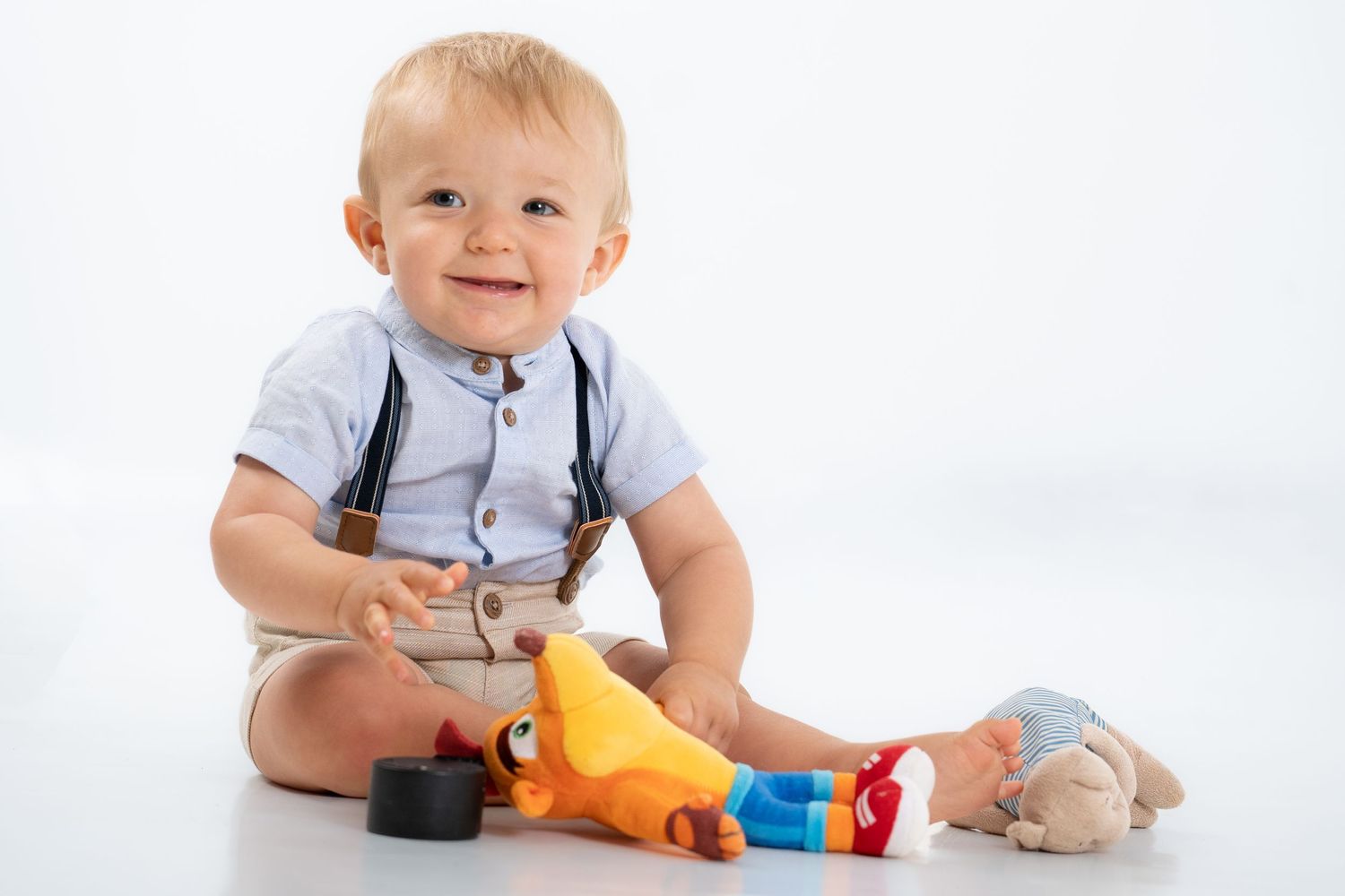 Smiling baby in formal outfit playing with colorful toys against white background.