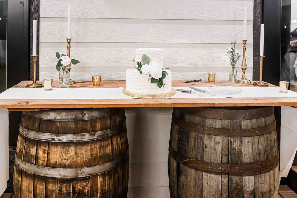 Rustic wedding bar setup with wooden whiskey barrels supporting a natural wood tabletop decorated with elegant flowers.