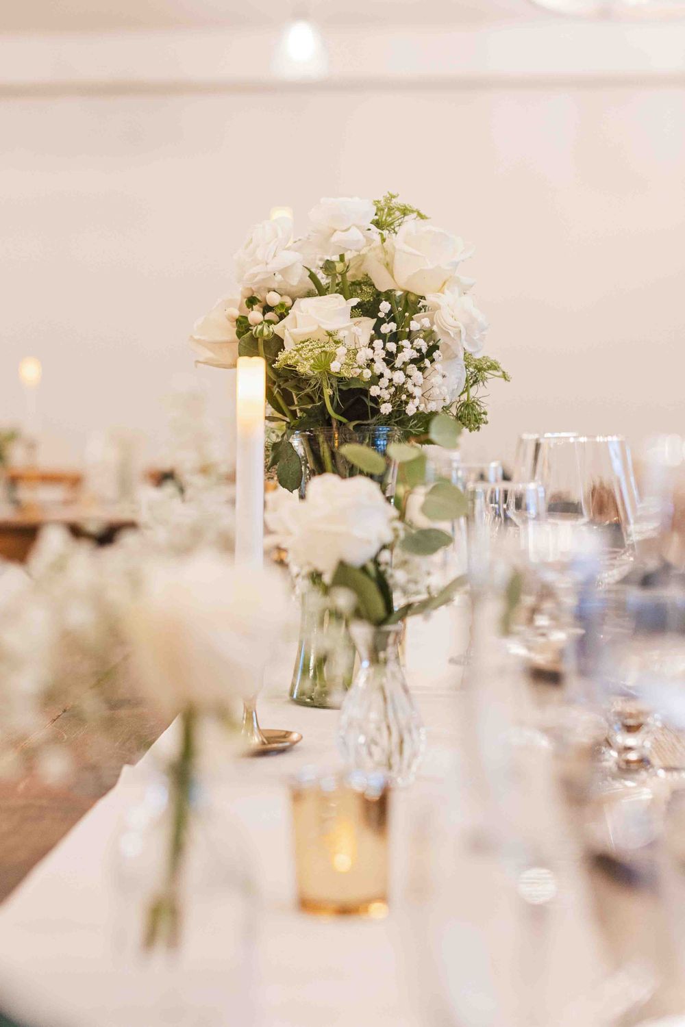 Elegant white rose and baby's breath floral centerpiece in glass vase on wedding reception table with wine glasses.