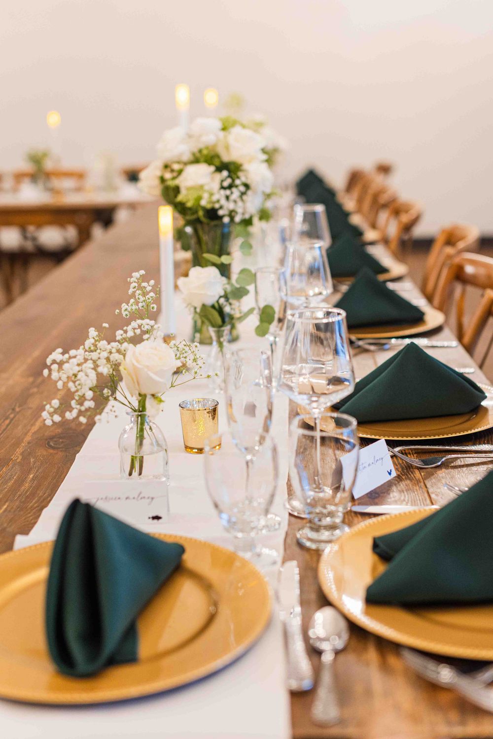 Elegant wedding table setting with gold charger plates, dark green napkins, and delicate white floral centerpieces.