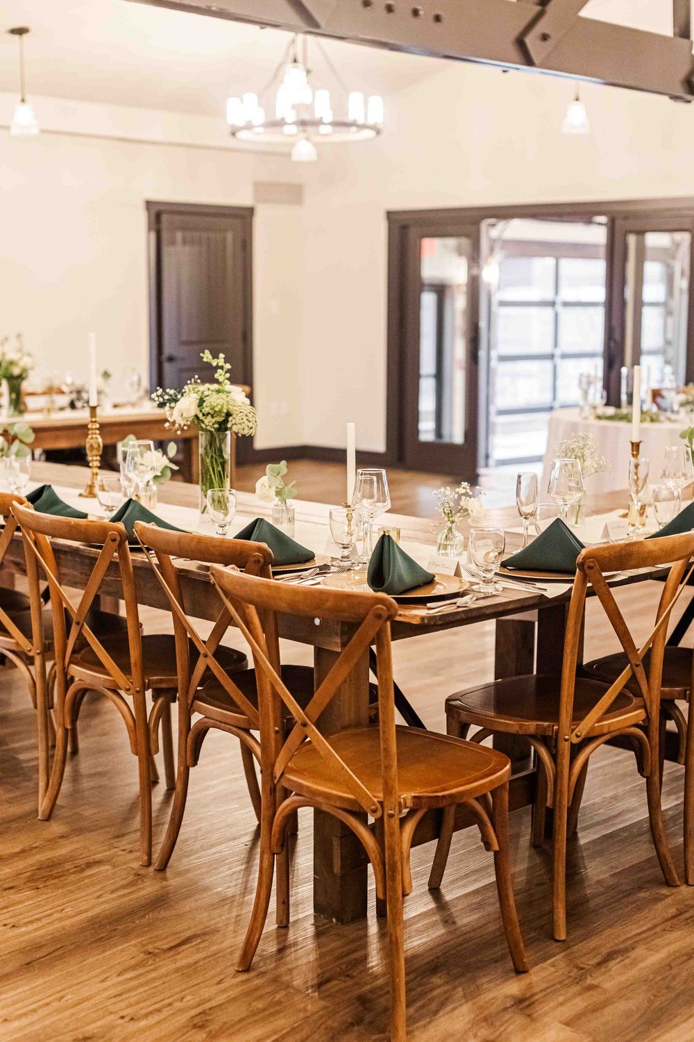 Elegant dining room setup with wooden crossback chairs, forest green napkins, and wine glasses on rustic farm tables.