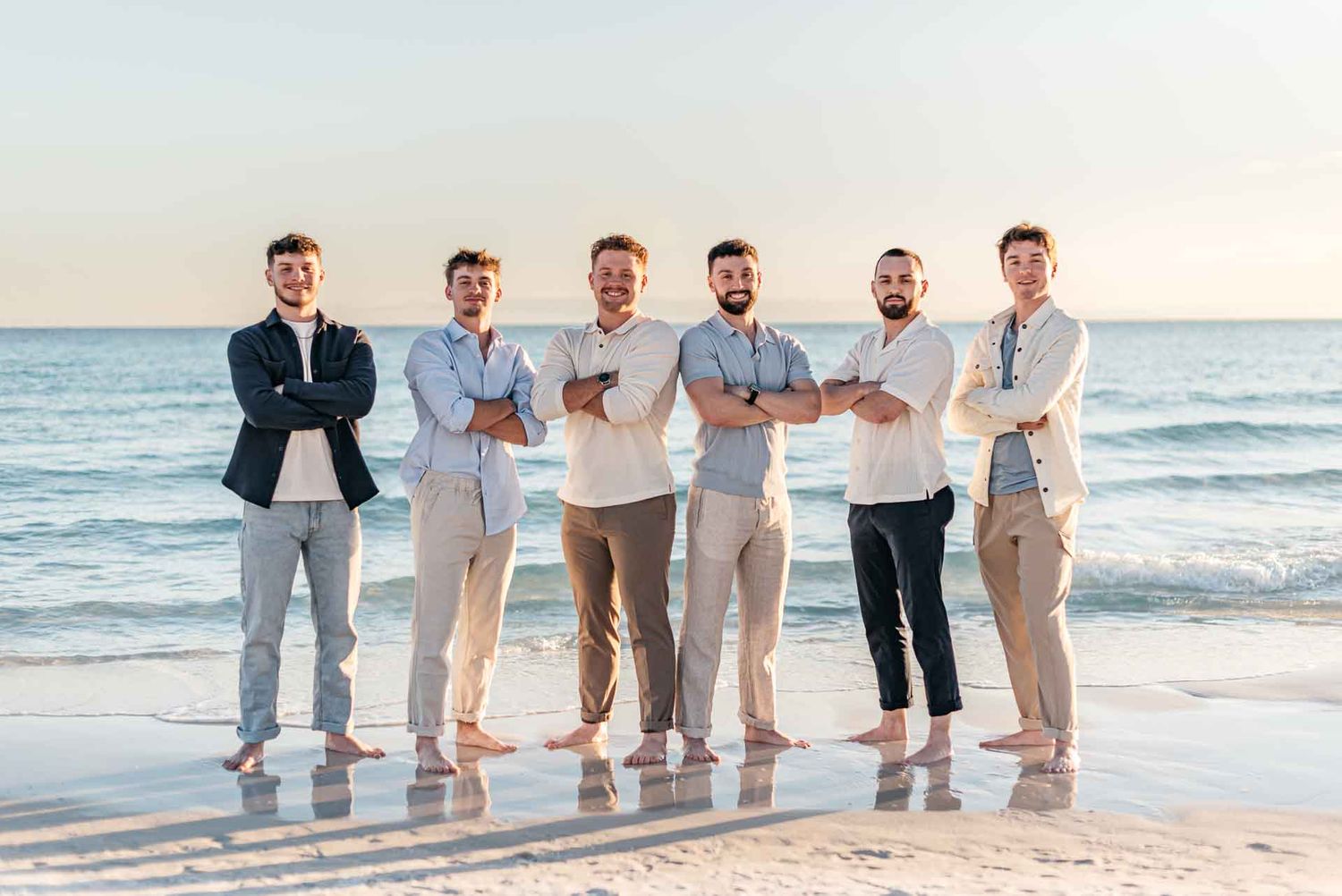 Row of people in coordinated beige and white outfits posed on beach at sunset.