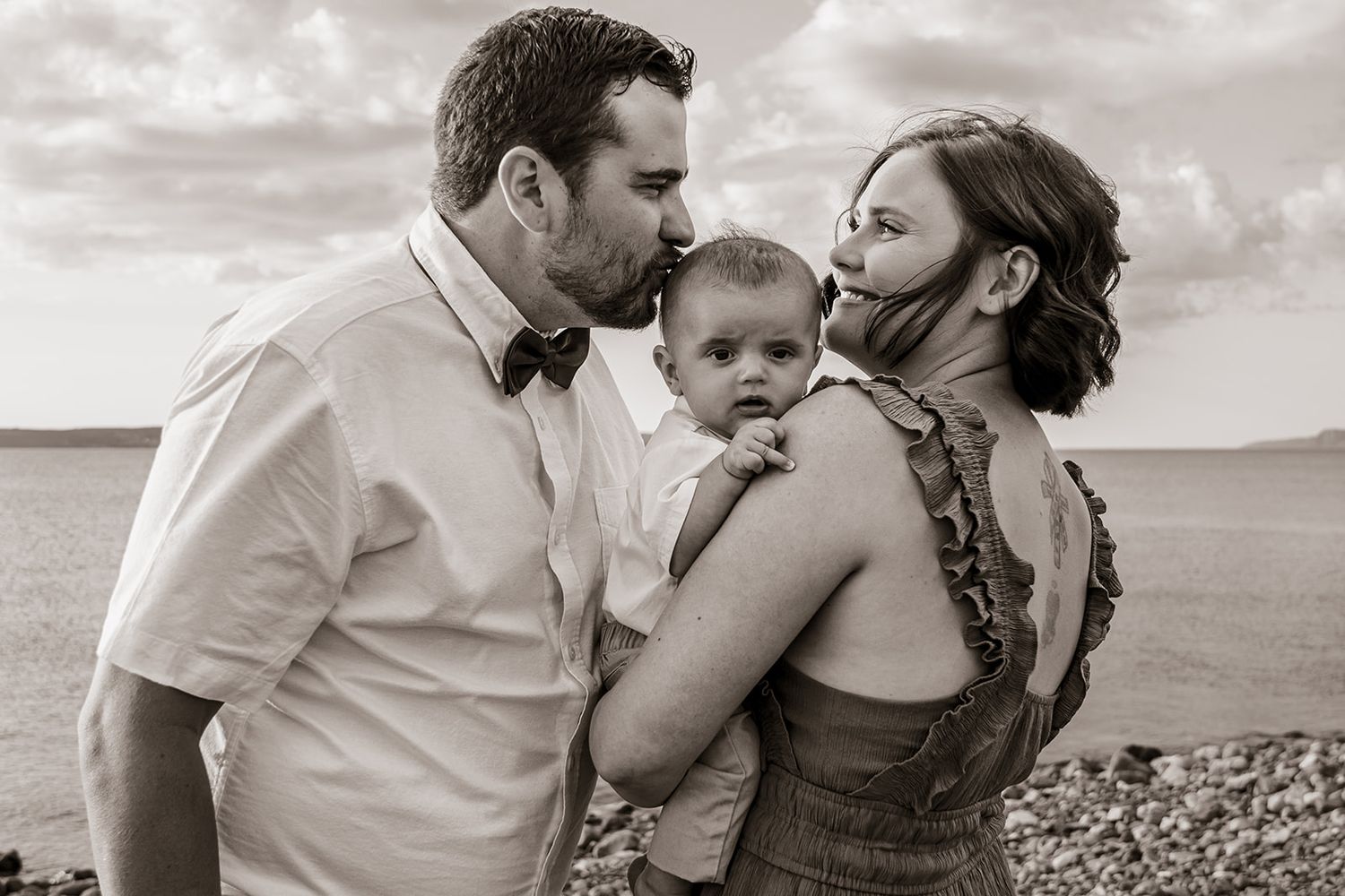 A black and white candid family portrait shows affectionate parents sharing a tender moment with their young child on a beach.