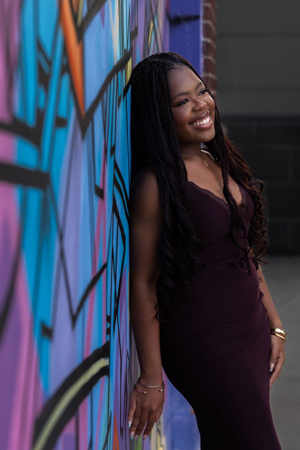 A smiling Black woman in a purple dress leans against a colorful graffiti wall with long braided hair.
