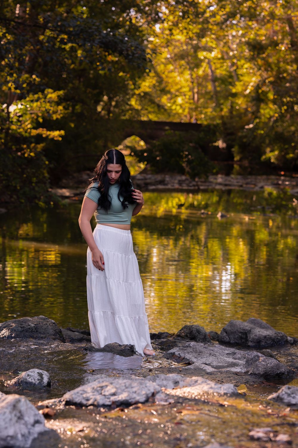 Young woman in white maxi skirt and green top standing on rocks by a reflective pond in a lush park.