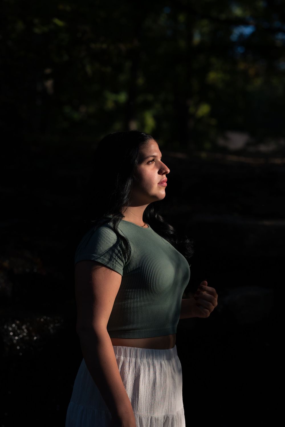 Young woman in green crop top and white skirt standing in dappled sunlight against dark forest background.