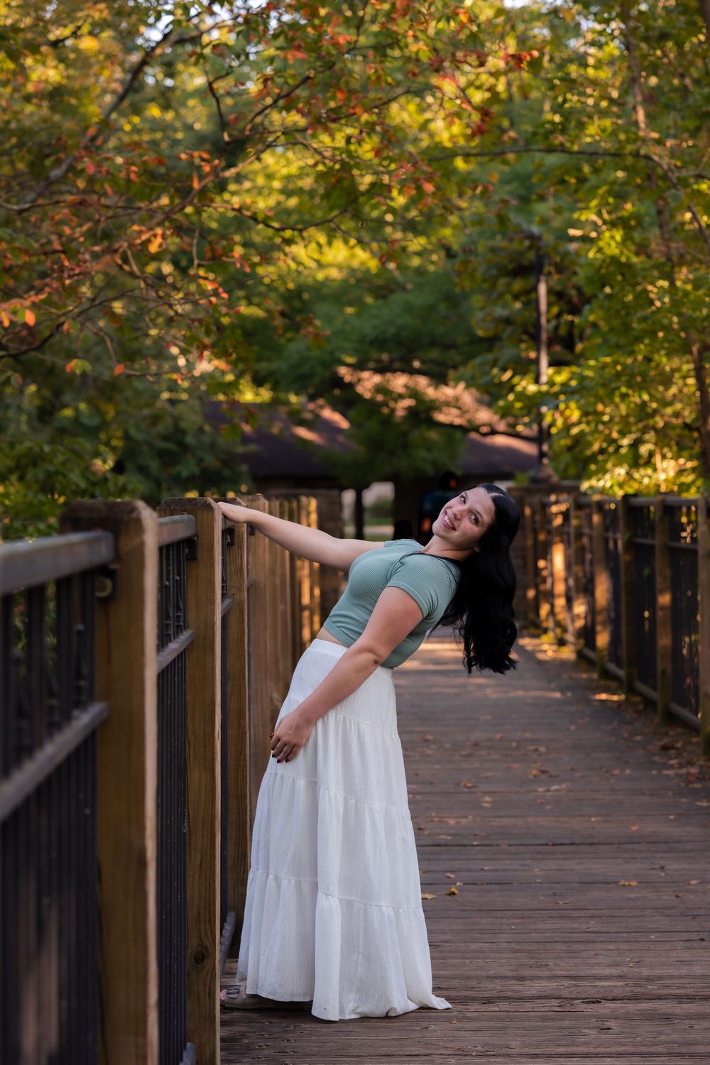A smiling woman in a white maxi skirt poses on a wooden bridge surrounded by autumn trees.