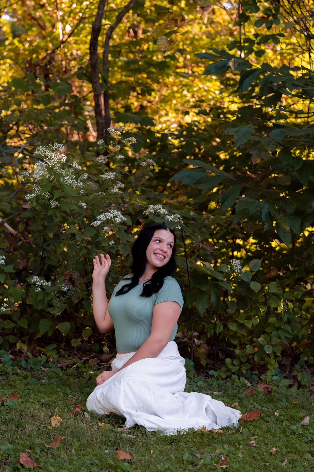 A smiling brunette woman in a green top and white skirt sits on grass beside blooming white wildflowers in a lush garden.