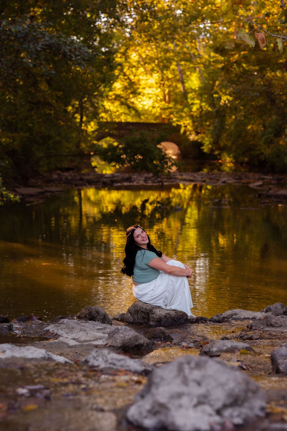 A woman in a white skirt sits on rocks by a golden autumn creek, smiling joyfully with trees reflected in the water.
