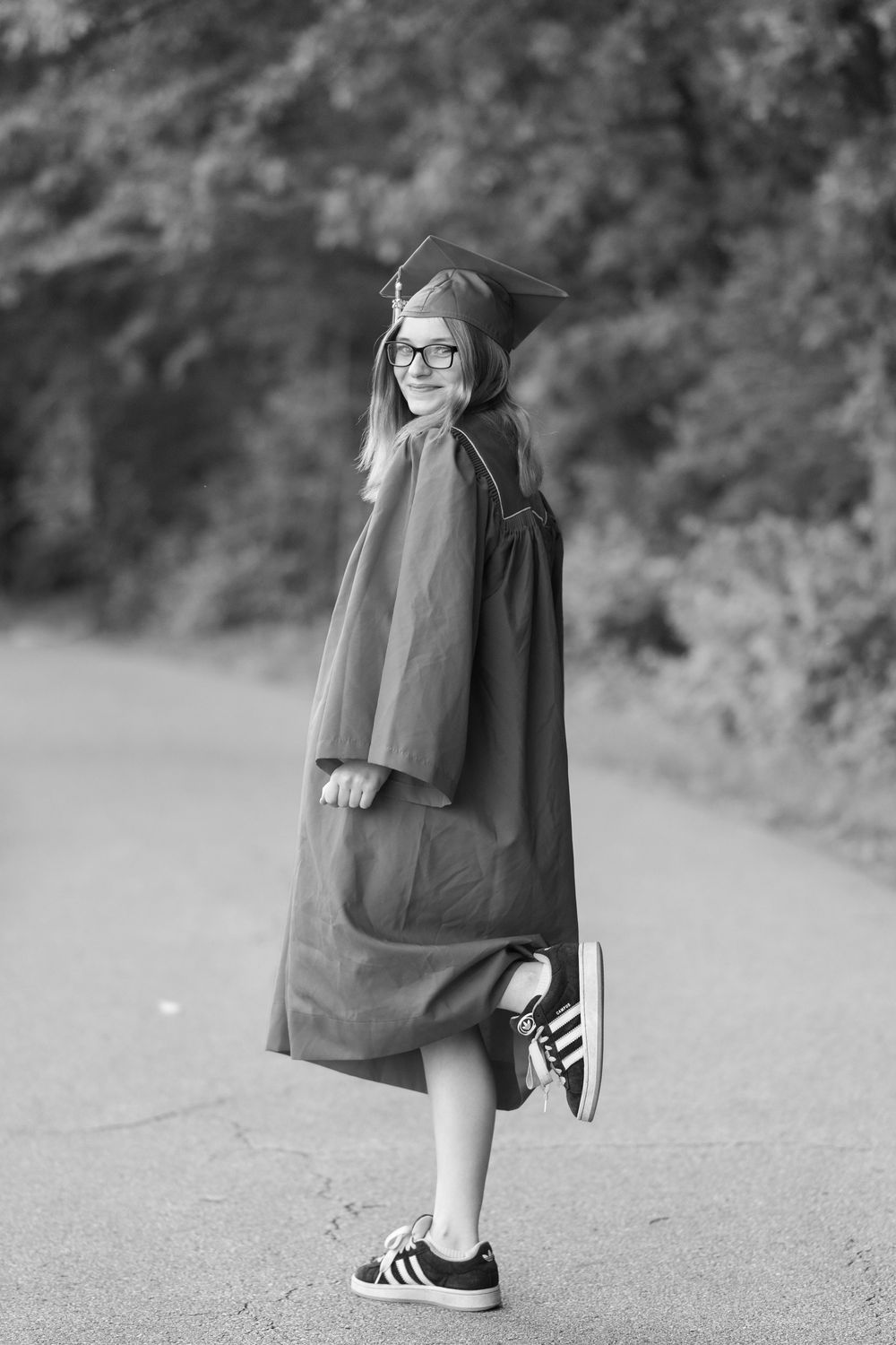 Black and white portrait of graduate in cap and gown wearing casual sneakers on a path outdoors.