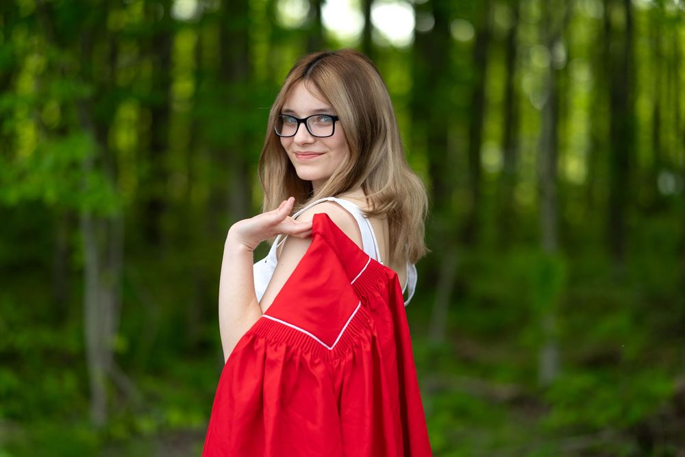 Someone in glasses stands in a forest setting holding a bright red dress against a green background.