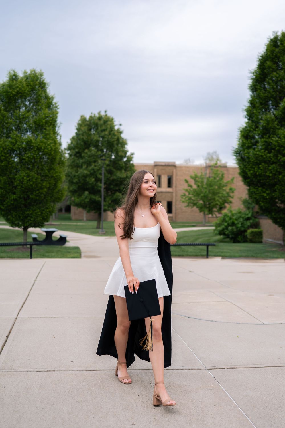 Young woman in white dress holding graduation cap walks on campus path with green trees and brick building behind her.