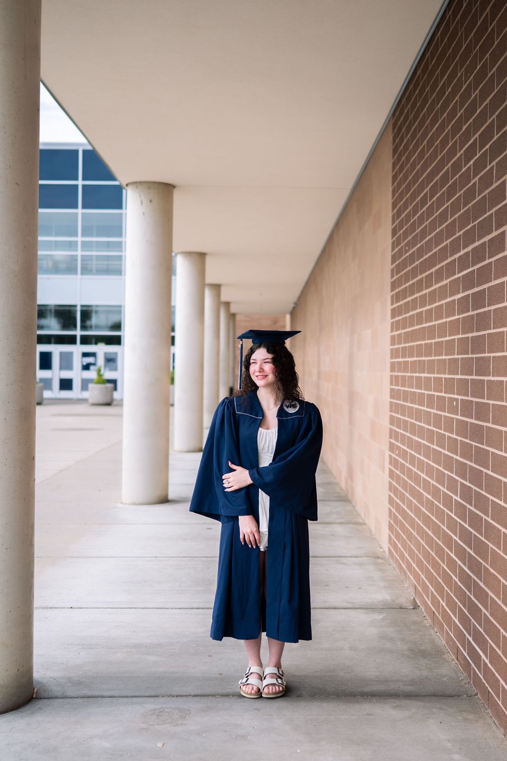 A young woman in navy graduation gown and cap smiles while standing in a columned walkway beside a brick building.