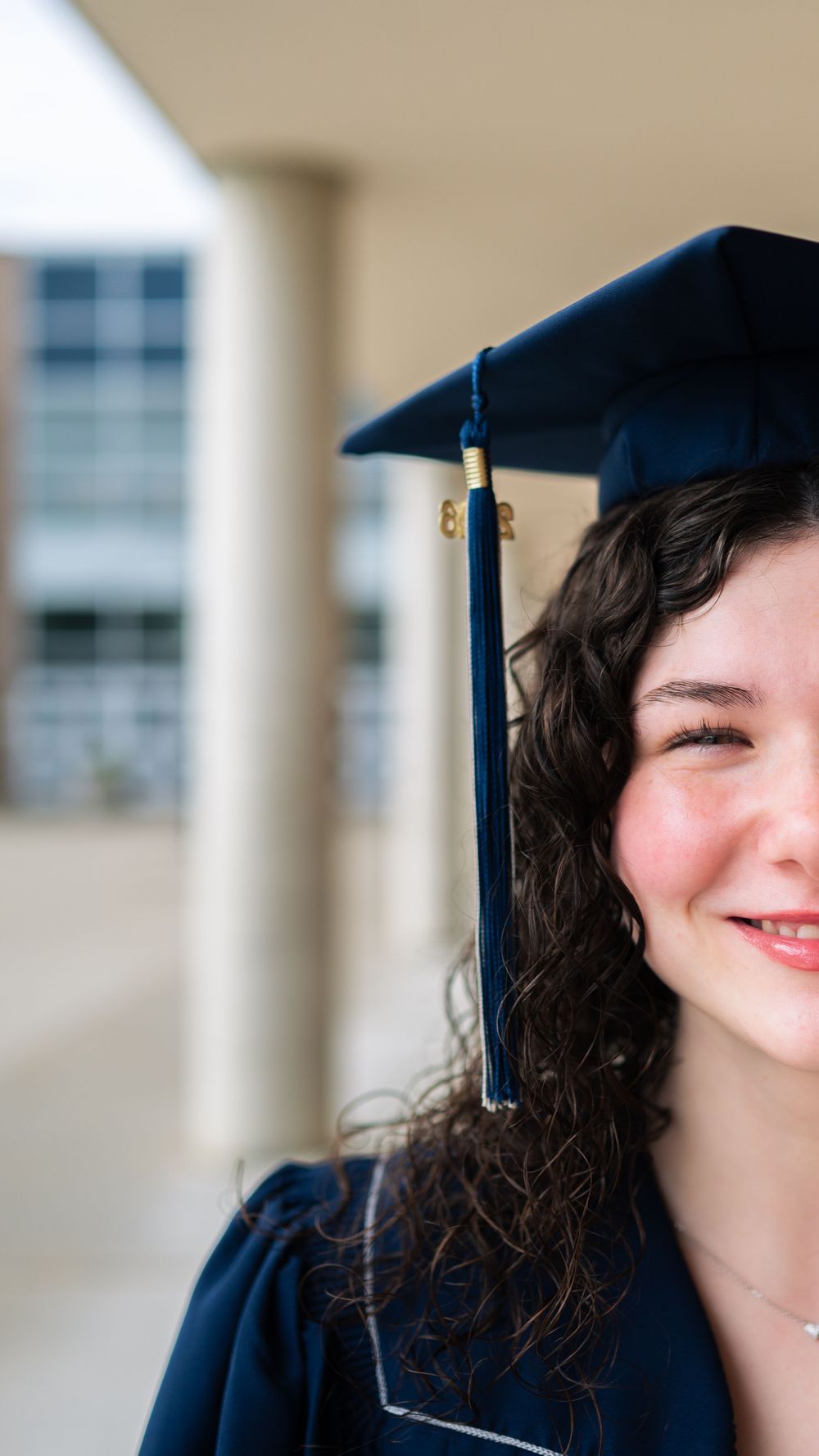 Smiling young woman in navy blue graduation cap and gown with curly hair, celebrating academic achievement outdoors.