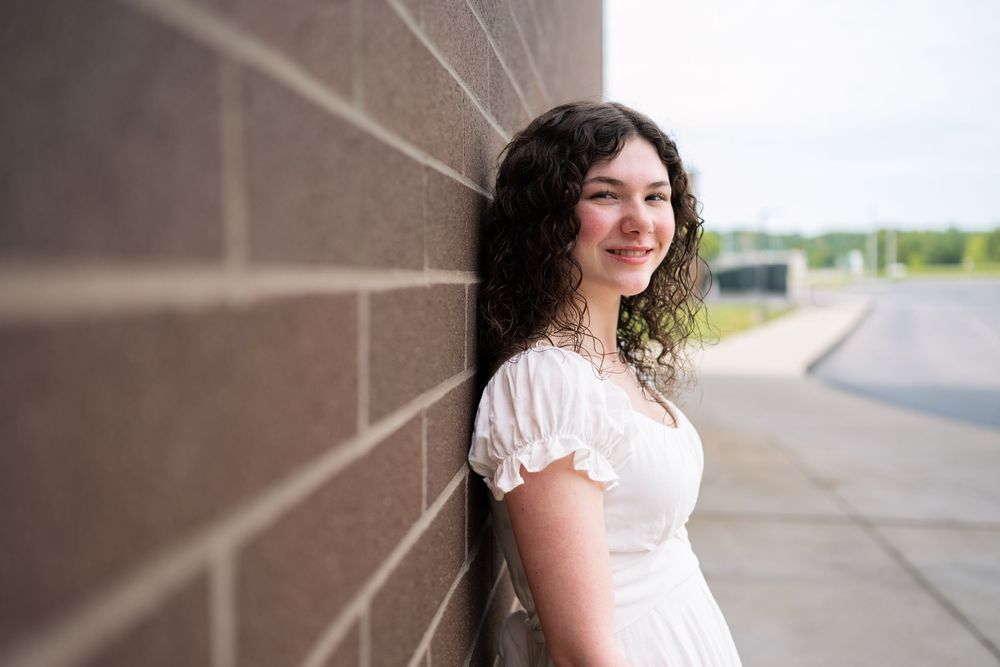 Young woman with curly dark hair in white dress smiling against brick wall outdoors.