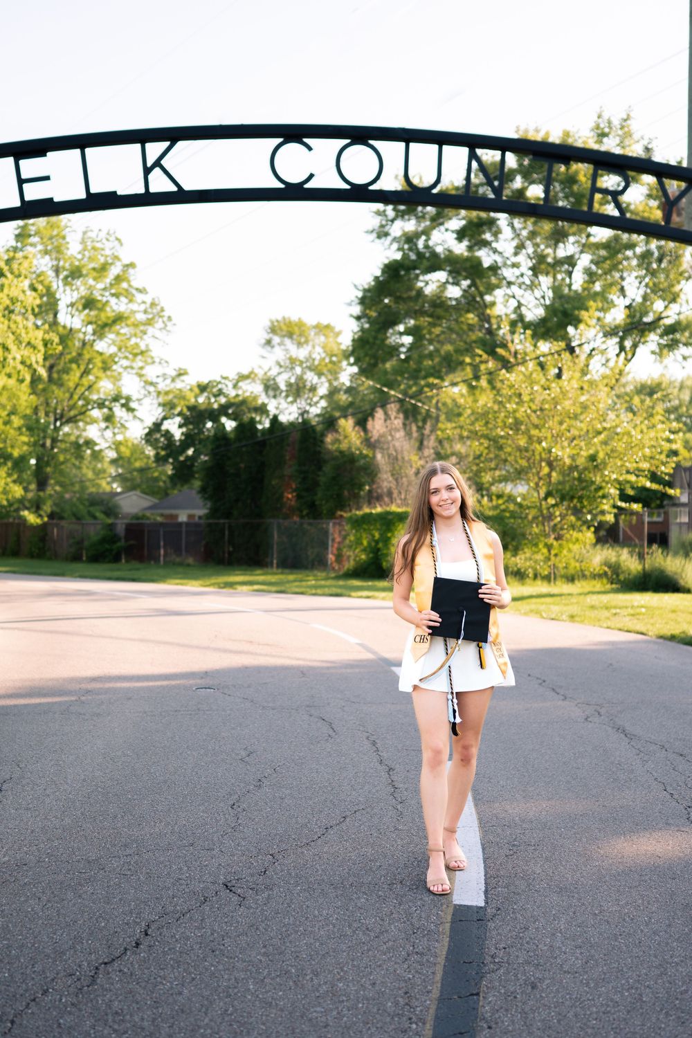 Young woman in white dress holds diploma under Elk Country arch sign on sunny day.