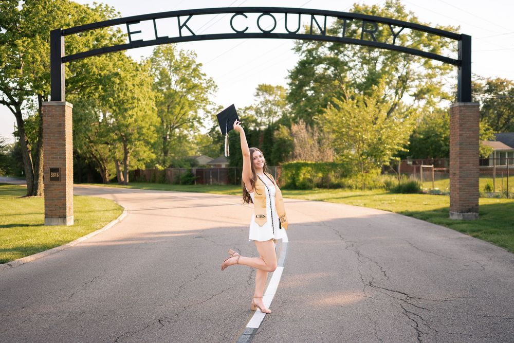 Graduate in white gown joyfully leaps under Elk County arch, tossing cap in the air on a sunny day.