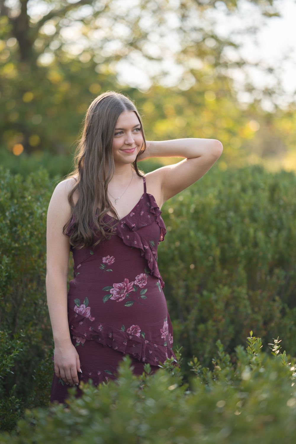 A person in a purple floral dress poses outdoors among green foliage during golden hour.