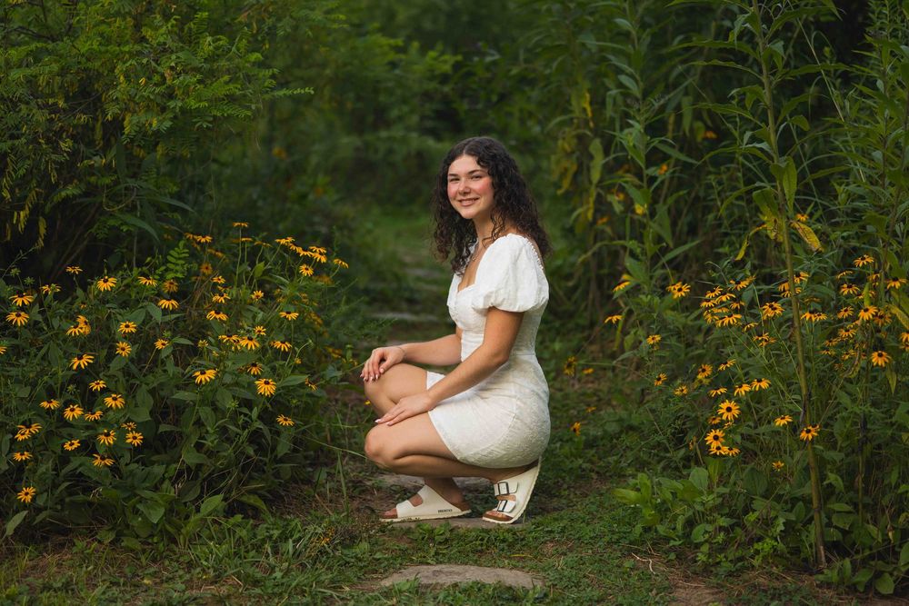A person in a white dress crouches in a garden filled with yellow wildflowers on a sunny summer day.