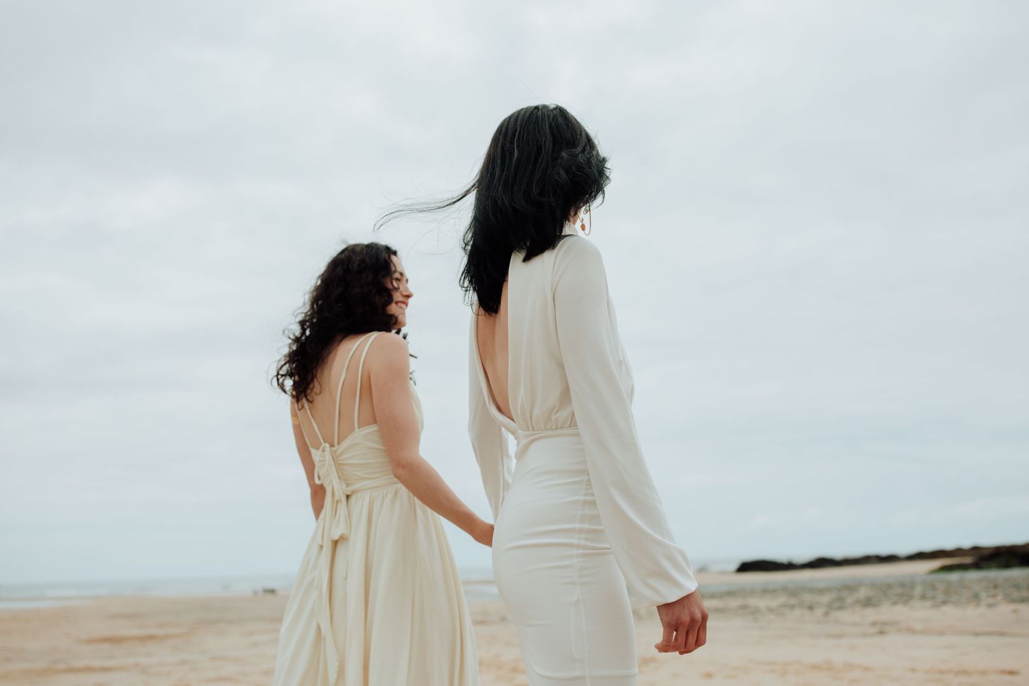 Minimalist beach wedding portrait of two people in white dresses against cloudy sky. Cornish wedding elopement