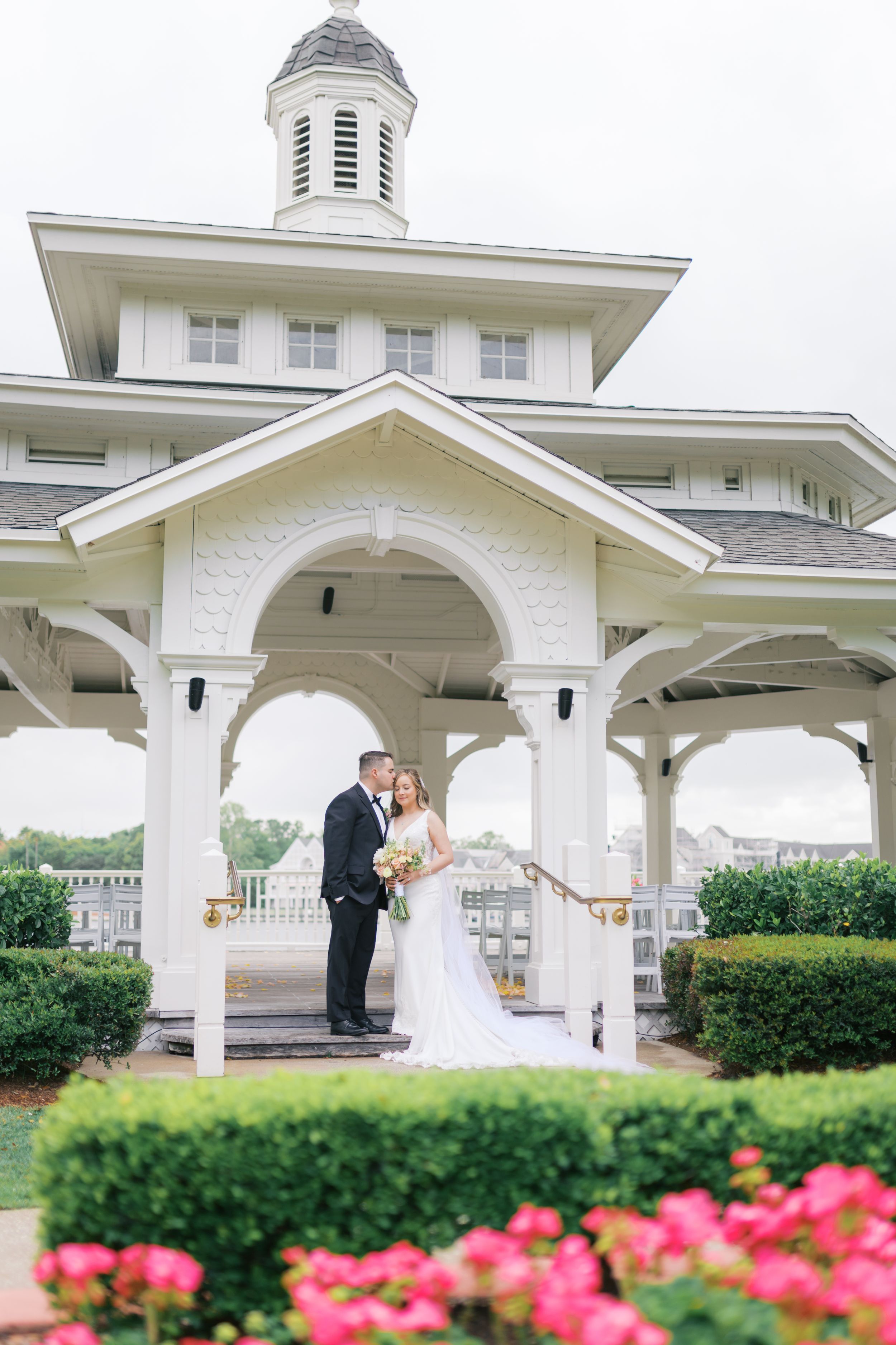 Bride and groom pose under elegant white gazebo with pink flowers in foreground at outdoor wedding venue.
