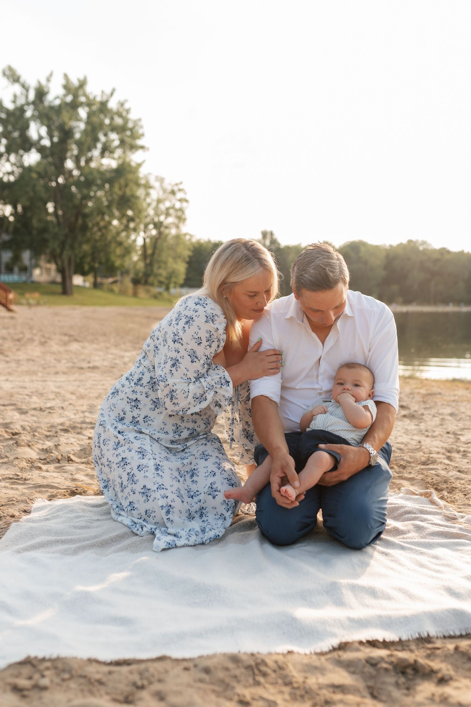 A family sits on a blanket on a sandy beach, with parents lovingly attending to their baby in golden sunlight.