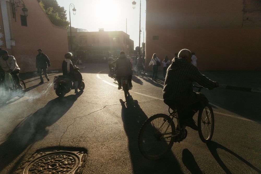 Cyclists and scooters ride through a sunlit urban street at golden hour, casting long shadows on the pavement.
