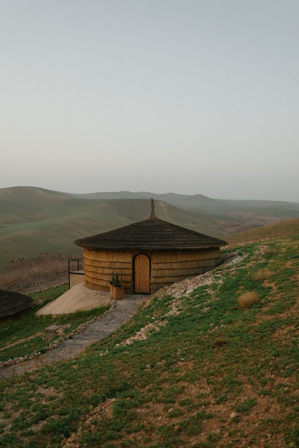 A rustic circular mud hut with a conical roof sits on a green hillside at dusk with rolling desert mountains behind.