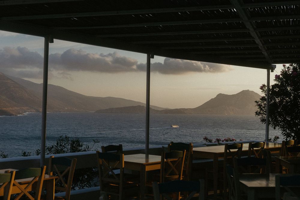 Sunset view from a seaside restaurant terrace with wooden chairs, tables, and mountain silhouettes over calm water.