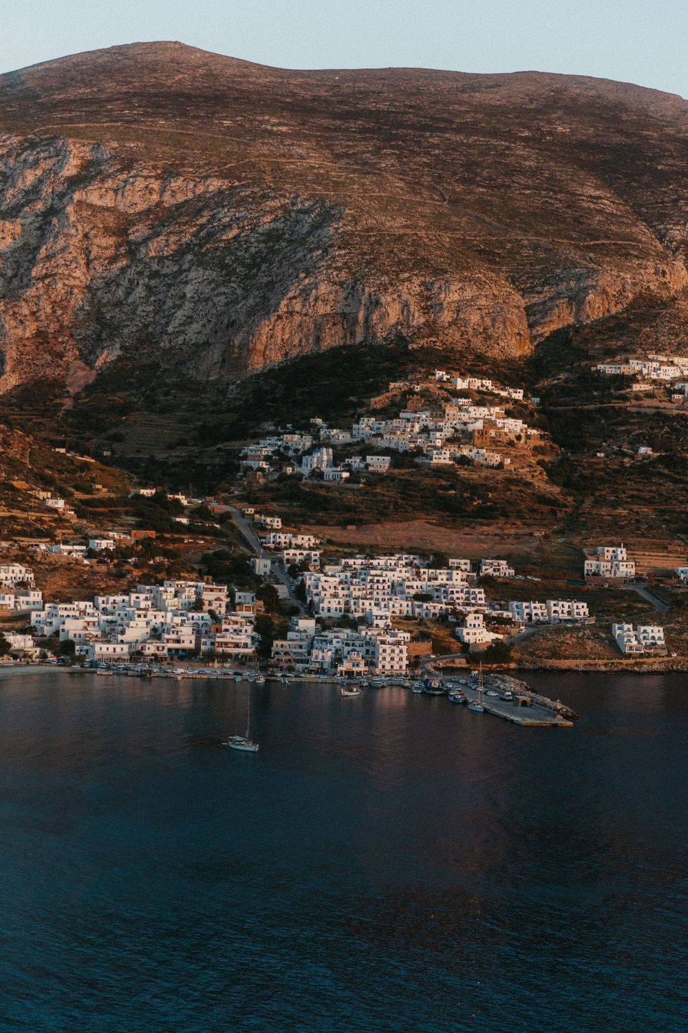 Aerial view of a white-washed Greek village nestled on a rocky hillside overlooking a calm blue sea at golden hour.