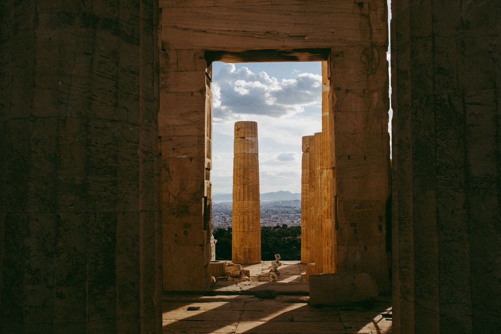 Ancient Greek columns framed by stone gateway at the Acropolis Athens, with city skyline and dramatic sky beyond.