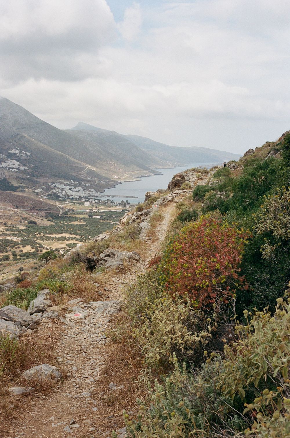 A narrow dirt hiking trail winds down a rocky hillside overlooking a coastal bay and village in Greece.