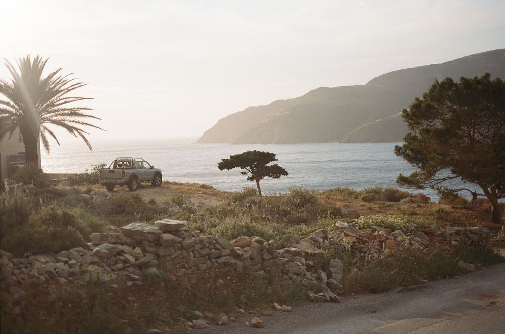 A silver SUV parked near a rocky Mediterranean coastline with palm trees, lush greenery, and misty mountain views.
