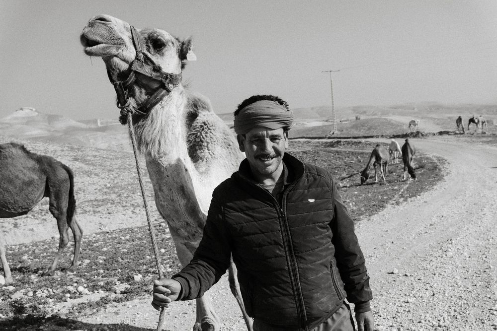 Black and white photo of a smiling man leading a camel along a rural dirt road in an arid landscape.