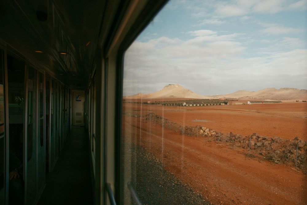 View from a train window showing red desert landscape and distant mountains under blue sky.