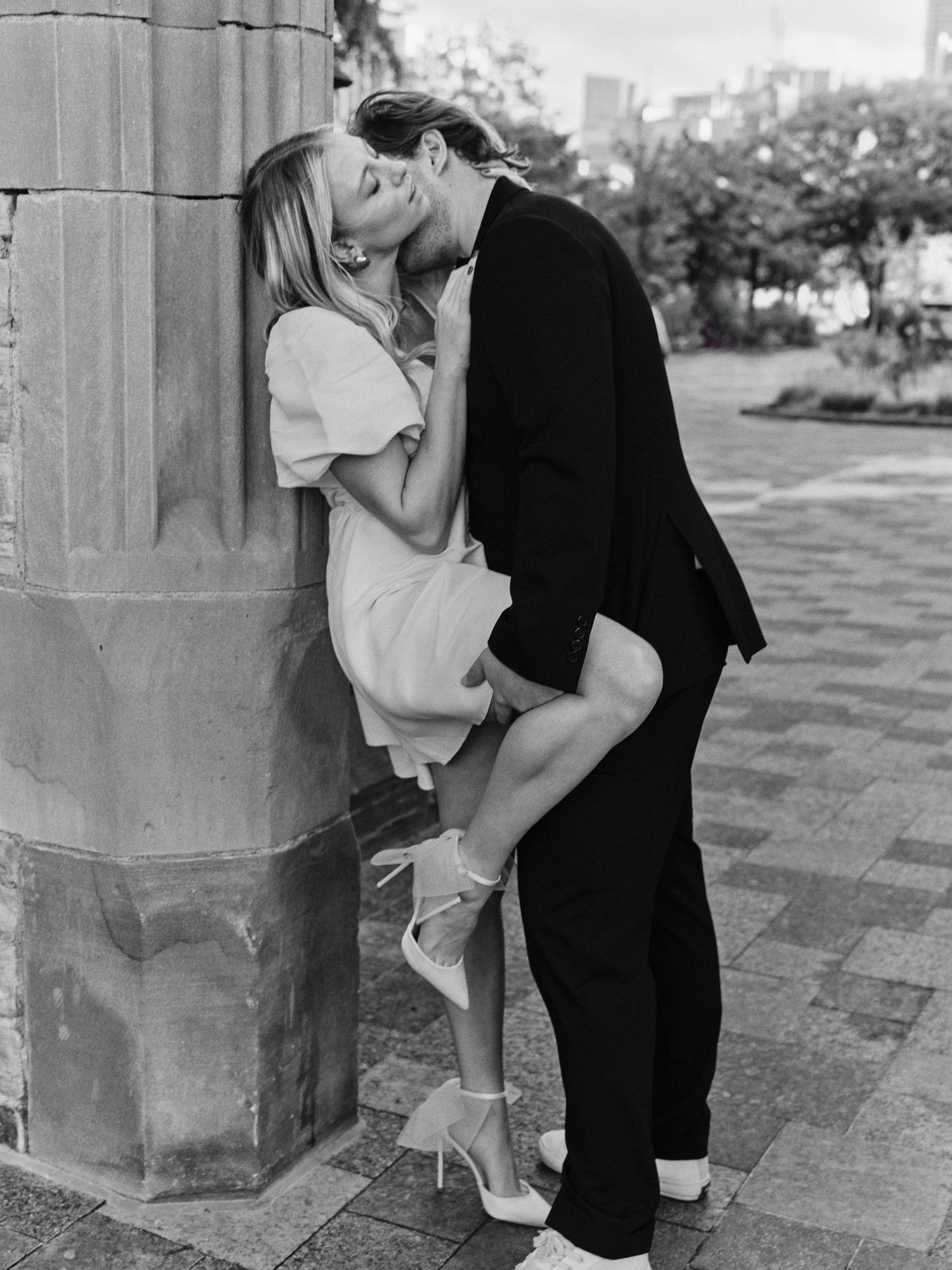 A romantic black and white photograph of a couple embracing against a stone pillar outdoors.