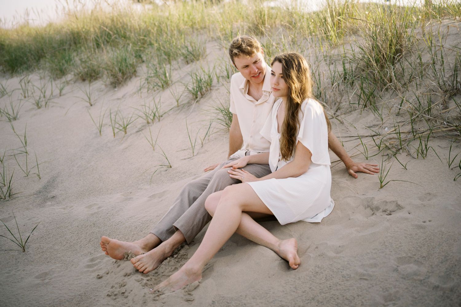 Two people in white outfits sit together on sandy beach dunes during a warm summer evening.