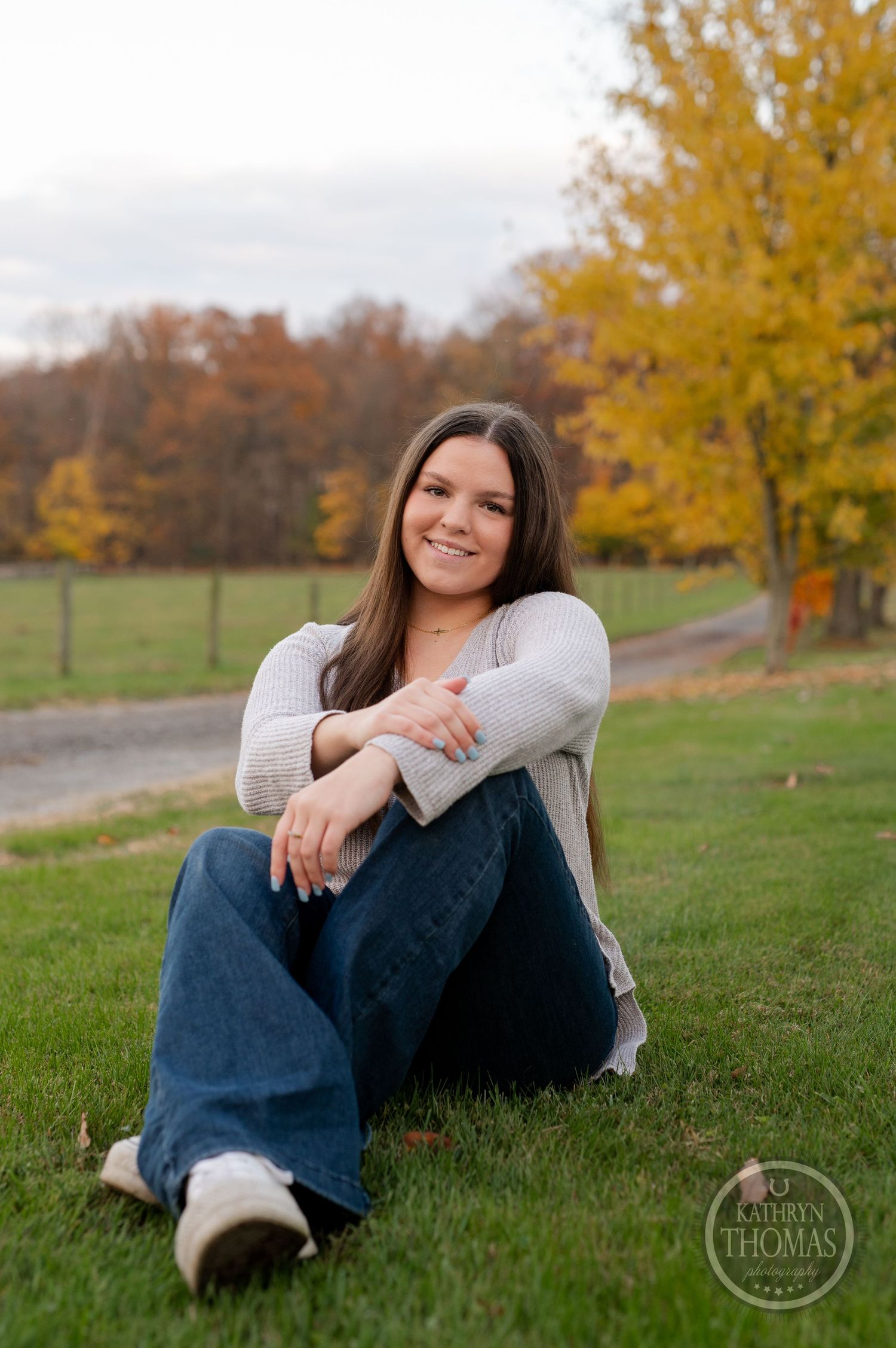 Person in gray sweater and jeans sits on grass with vibrant fall foliage in background.